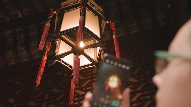 Temple lantern glowing under wooden roof, visitor with glasses captures warm light on red tassels using smartphone, carved ceiling patterns visible, intimate evening ambience with cultural heritage