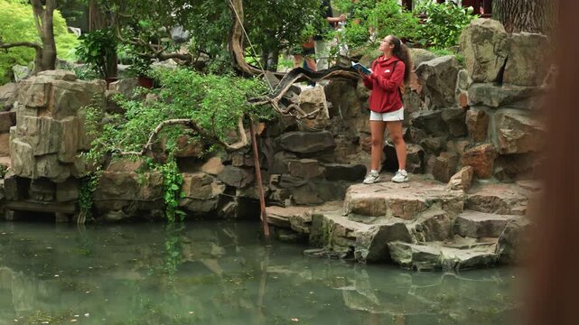 Caucasian woman feeding koi pond playful moment on stone steps near lush rockery red jacket, sneakers, throwing breadcrumbs vlogger, birdwatcher, urban explorer roles splashes, water ripples, ambient