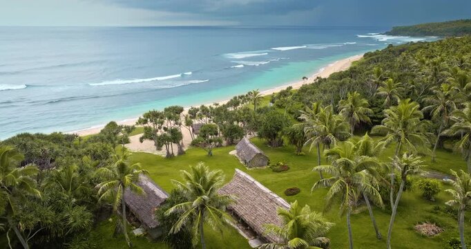 Port Resolution beach on Tana Island in Vanuatu, presenting a beautiful coastline with white sand, turquoise water, breaking waves, palm trees, and traditional thatched huts. Zoom out aerial flight