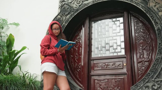 Standing reader by carved circular gate, hooded red jacket and white shorts, leaning against carved wooden moon gate, tropical plants at feet, attentive gaze flipping turquoise notebook pages, playful