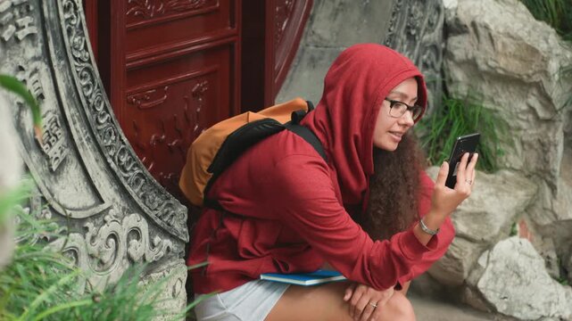 Asian woman red hoodie checking phone, southeast asian student traveler sits by ornate circular temple gate, backpack and glasses, scrolling map on phone, contemplative expression, mossy stone, green