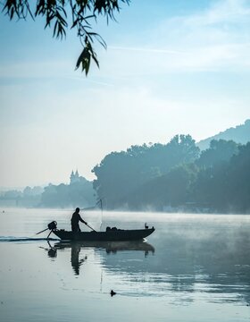 Serene Morning on the River - A Lone Fisherman Navigates Misty Waters with Distant Hills and Overhanging Branches.