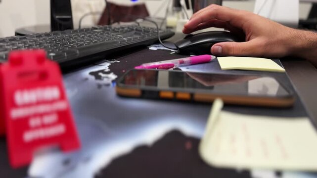 hand using a computer mouse at an office desk with keyboard, smartphone, sticky notes, pink pen, and a blurred red warning sign in the foreground.