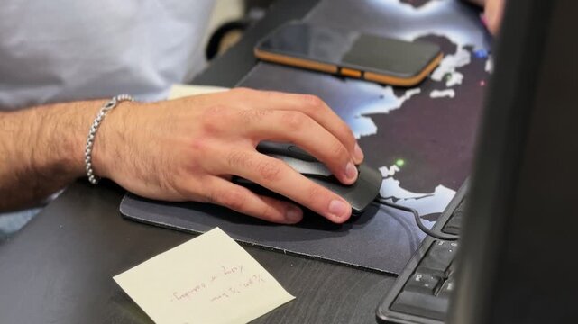 Close-up of a hand using a computer mouse at an office desk with keyboard, smartphone, and sticky notes, showing focused desk work in an indoor workspace setting.