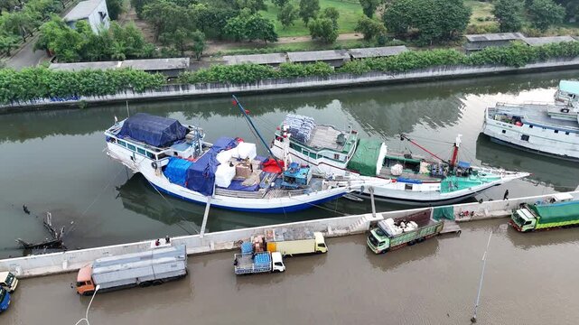 Aerial view of traditional wooden Pinisi cargo ships and colorful vessels docked at Sunda Kelapa historical port with stacks of shipping containers and Jakarta cityscape in the background.