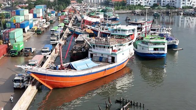 Aerial view of traditional wooden Pinisi cargo ships and colorful vessels docked at Sunda Kelapa historical port with stacks of shipping containers and Jakarta cityscape in the background.