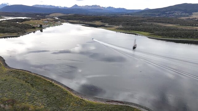 Cinematic flies forward past the anchored Schooner Amazone, tracking a fast speedboat as it cuts a long white wake through the calm waters of Bahia Harberton, Argentina