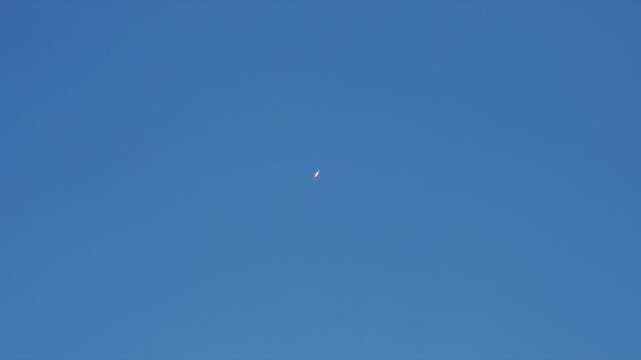 Rocket with bright orange flame ascends diagonally through clear blue sky leaving long white exhaust vapor trail behind. Space mission liftoff captured from ground with growing contrail