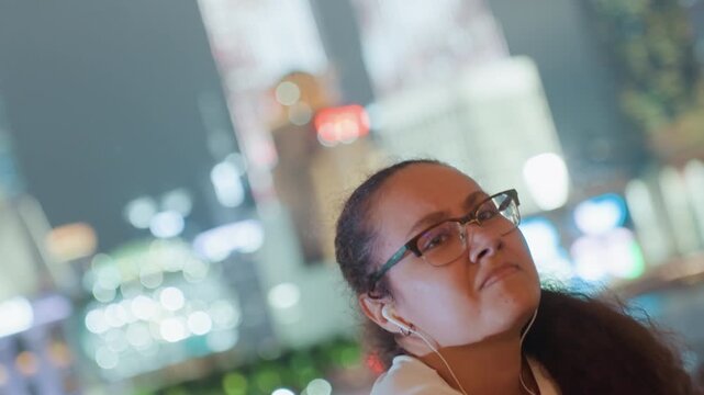 Young woman dancing under city lights, waterfront skyline bokeh, earbuds in, shifting from calm to exuberant, arms raised, candid joyful expression, neon reflections, urban nightlife energy.