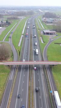 Vertical screen Aerial Motorway Container Trucks Crossing Under Pedestrian Bridge, Multiple Trailers And Box Trucks In Motion, Roadside Service Area, Green Margins And Steady Logistics Tempo