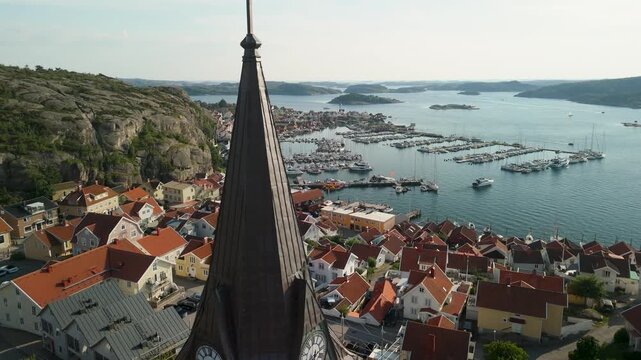 Drone circles Fj&auml;llbacka church steeple to reveal red-roofed town, granite cliffs, packed marina and scenic Bohusl&auml;n archipelago in warm summer evening light
