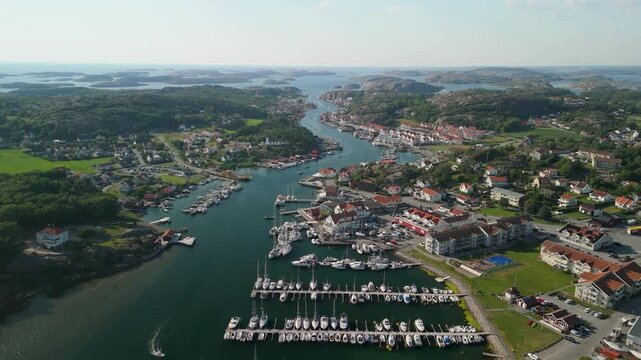 Aerial orbiting above Fiskeb&auml;ckskil marina on Skaft&ouml; island showing sailboats docked in crystal blue water surrounded by coastal village houses and rocky islands in Swedish archipelago
