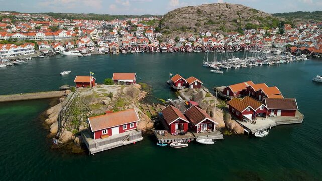 Drone parallax circles Falun red cottages on granite islets in Hunnebostrand harbor, Bohusl&auml;n, with sailboats, Swedish flags and coastal town in background