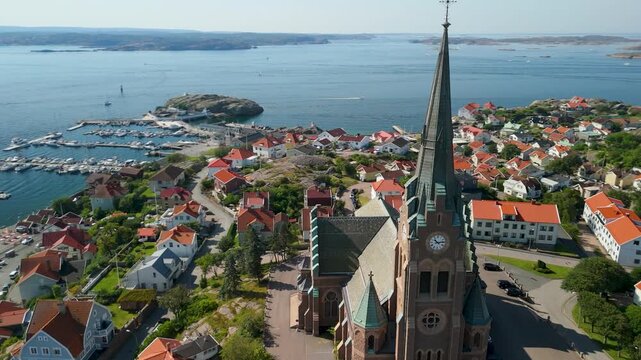 Aerial drone orbits Lysekil Church clock tower revealing Swedish coastal cityscape of orange rooftops granite hills harbor and sparkling sea beyond
