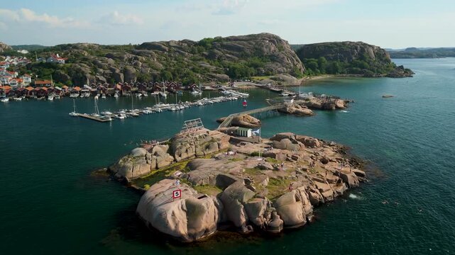 Aerial view of Bovallstrand marina and granite islet in Bohusl&auml;n Sweden with tourists sunbathing swimming and diving while sailboats fill harbor beyond