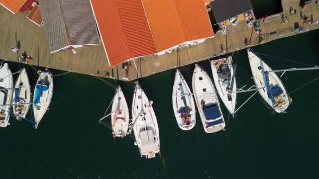 Top down aerial sliding along Sm&ouml;gen marina boardwalk in Sweden with white sailboats moored perpendicular to pier and tourists strolling in summer