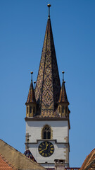 Gothic steeple of the Lutheran Evangelical Cathedral in Sibiu, Romania, featuring colorful glazed roof tiles and clock tower under a blue sky. © Dan
