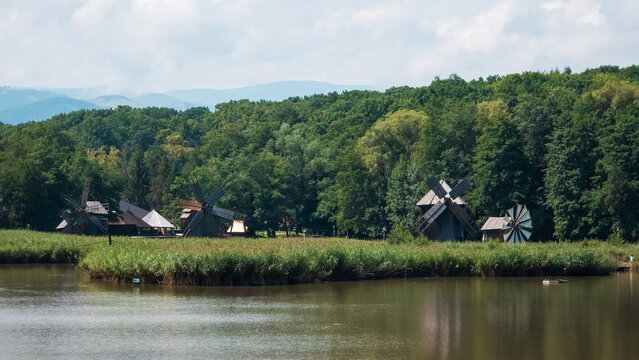 Panoramic view of traditional wooden windmills lined along the lake shore at ASTRA Museum in Sibiu, Romania. Scenic landscape with lush forest and mountain backdrop under summer clouds