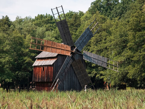 Traditional wooden windmill with large wooden sails at the ASTRA Museum in Sibiu, Romania. Medium shot of rustic rural architecture set against dense green forest trees in daylight.