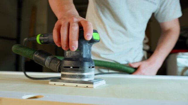 Worker sanding wooden cabinet surface with an electric orbital sander during renovation. Interior carpentry process preparing furniture for finishing. Close up view
