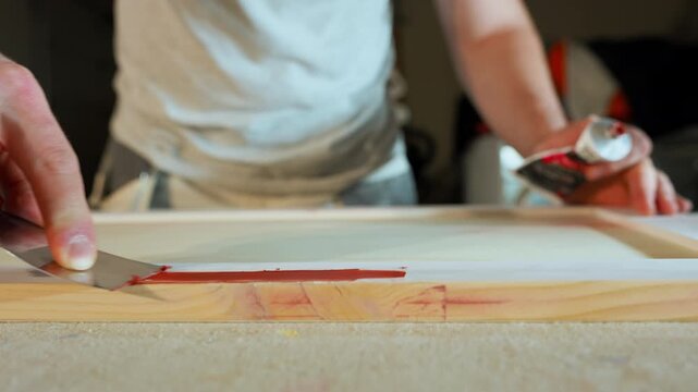 Worker applying wood filler to cabinet frame with a spatula during furniture production. Close-up of carpentry process preparing wooden surface for sanding and finishing in cabinetry manufacturing