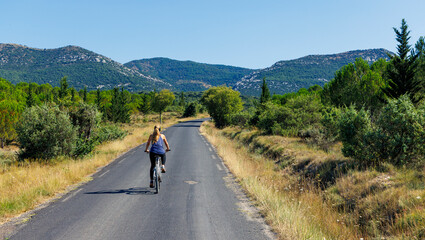 Obraz premium Young woman riding a bicycle on a scenic empty road in Provence, France, with mountains and pine trees, sustainable travel and freedom concept.