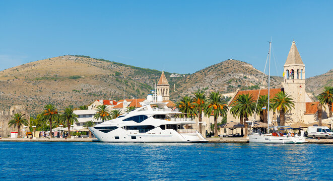 Luxury white superyacht docked at the historic waterfront of Trogir, Croatia, featuring the Cathedral of St. Lawrence and palm trees under a clear blue sky.