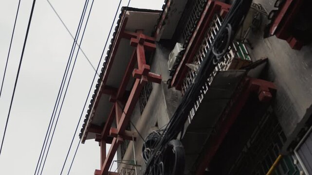 Overcast alleyway facade with wires moody closeup of traditional chinese building eaves and bundled utility cables, dark red wooden brackets and tiled roof, gritty concrete walls, leaning power pole