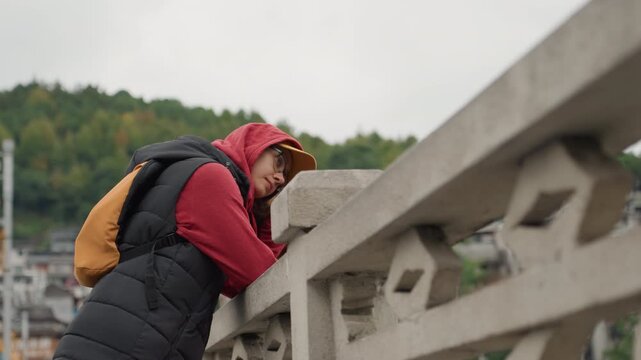 Woman leaning on ornate bridge railing near hillside curious explorer in red hoodie inspects stone details wearing cap backpack and glasses atmospheric overcast light highlights concrete texture