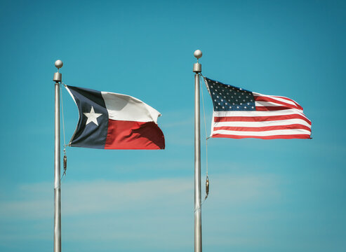 Texas state flag and American flag waving in the wind on flagpoles. Blue sky background. 