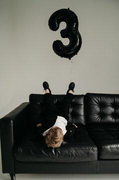 A little restless boy in a black suit plays on a black leather sofa on his birthday against the background of an inflatable balloon in the shape of the number three in a white photo studio