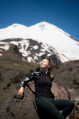 Fototapeta premium Female tourist chilling in the snowy mountains during a rest stop on a hike, resting with her eyes closed on the stone. A summer outdoor adventure. 