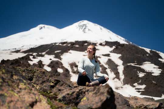 Young woman tourist sitting on a rock with snow-capped mountains and a blue sky in the background, in sunlight. Summer trekking and acclimatization before the ascent.