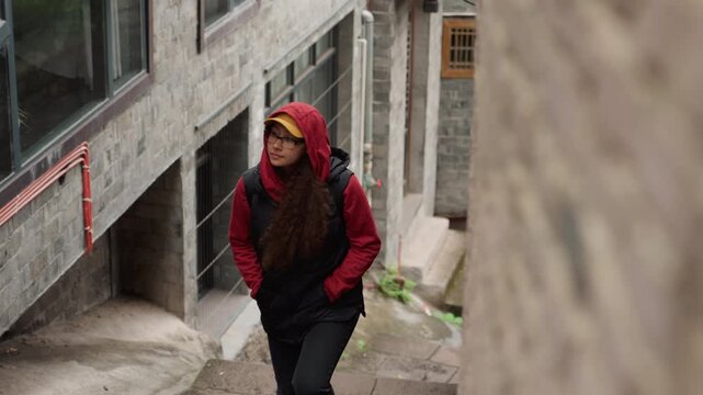 Red hoodie yellow cap gazing up narrow stone stairs and gray facades, long hair tucked inside hood, hands in pockets, thoughtful expression, urban architecture study, moody overcast light, candid