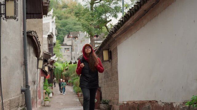 Young asian woman walking narrow alley in historic village, wearing red hooded jacket and backpack, cobblestone path lined with stone facades and lanterns, overcast autumn light, contemplative