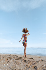 Young woman in colorful striped swimsuit walks barefoot on sandy beach near calm sea under clear...