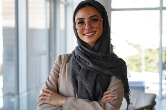 Smiling young woman in hijab and glasses poses confidently with arms crossed in a bright modern office
