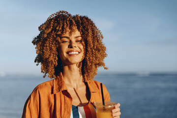 A happy woman with curly hair smiles enjoying a sunny day outdoors by the water while holding a...