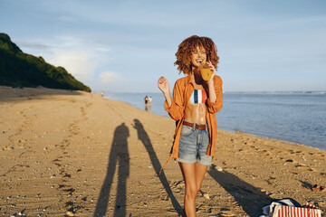 Happy woman with curly hair wearing shorts and open shirt drinks juice on sandy beach near ocean...