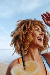 Happy woman with curly hair enjoys summer day at the beach holding cold drink with straw smiling...