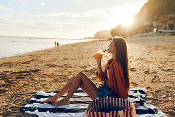 A woman in casual clothes sits on a striped towel on the beach drinking juice during sunset. The...