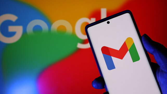 A hand holding a smartphone with the gmail app on the screen in front of a blurred google logo background with colorful shapes