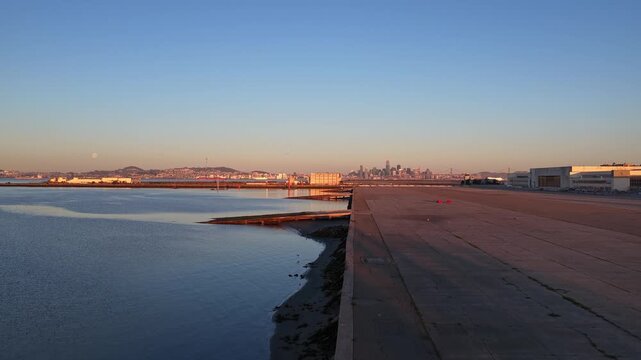 Aerial view flying over alameda waterfront park, a former naval air station runway, with the calm san francisco bay and the distant city skyline illuminated by the warm light of sunset