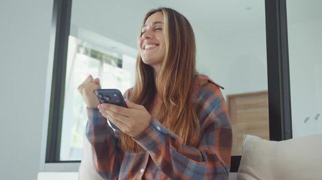happy woman reading message on smartphone at home smiling and celebrating good news. happy woman raising fist with joy while using mobile phone in bright modern interior indoors