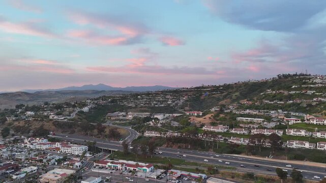 Aerial view of San Clemente at sunset with coastal hillside homes, freeway traffic in the foreground, and Saddleback Mountain visible in the distant background under pink evening clouds.