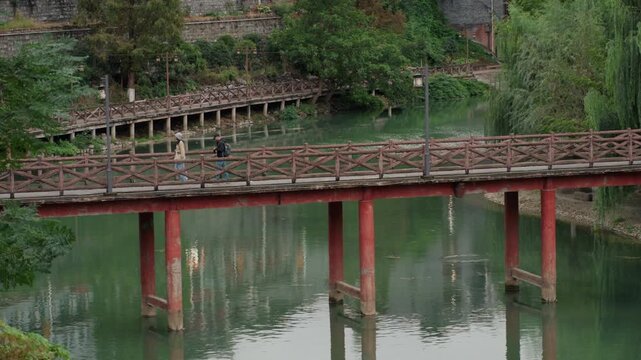 Red wooden bridge over green lake, local pedestrian strolling across historic railing, mossy pillars and calm water reflections, peaceful village backdrop, contemplative mood and scenic continuity