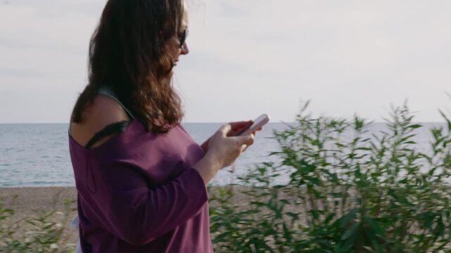 A woman walks along a seaside path, focused on her smartphone. The ocean and a sandy beach are visible in the background on a sunny day. Side view tracking shot