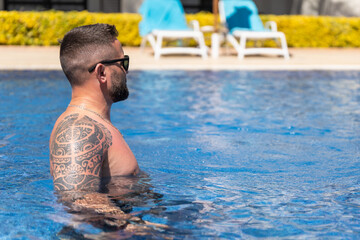 Side view of man with shoulder tattoo and sunglasses relaxing in blue swimming pool water at luxury...