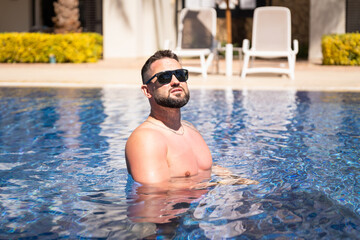 Handsome bearded man in sunglasses relaxing in swimming pool water at luxury tropical resort during...