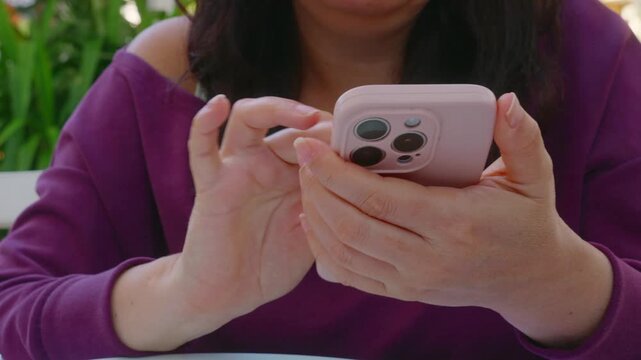 A woman in a purple sweater sits outdoors, holding a pink smartphone. Close up shot focuses on her hands as she types, scrolls, and texts on the mobile device